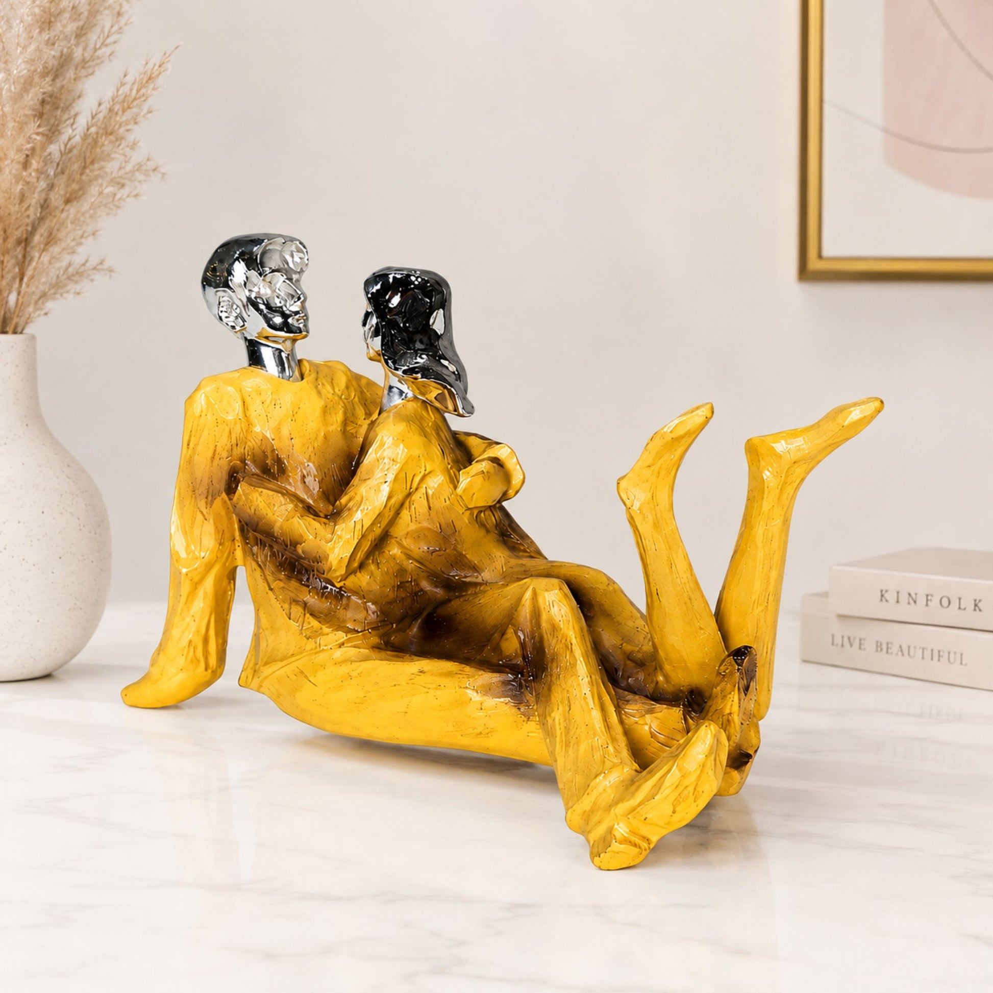 Abstract modern couple figurine with glossy chrome heads and textured bright yellow outfits reclining on a marble table.
