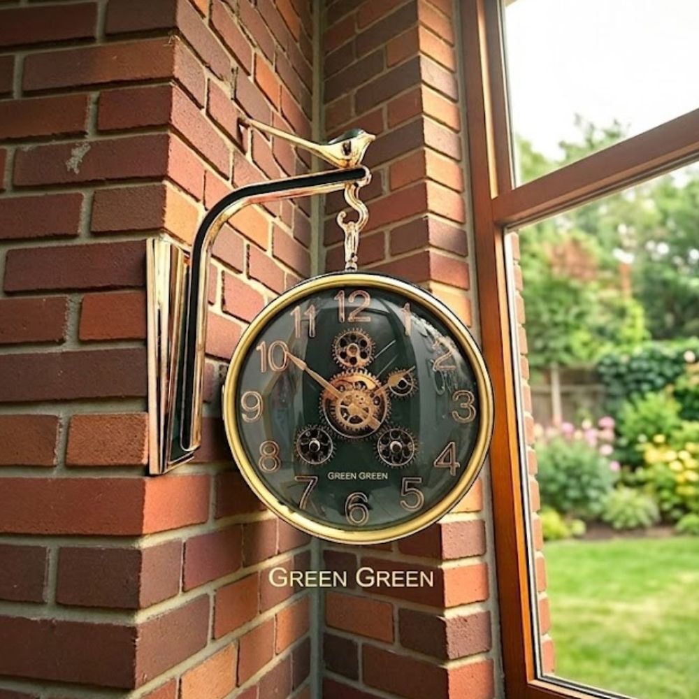 Double-sided hanging wall clock with a dark green dial, copper-gold numbers, and central gears, suspended from a polished gold bird bracket against a brick wall.