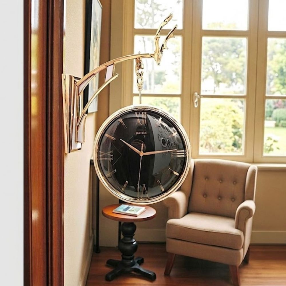 Double-sided hanging wall clock with a sleek black dial and gold Roman numerals, suspended from a polished gold deer antler bracket in a living room interior.