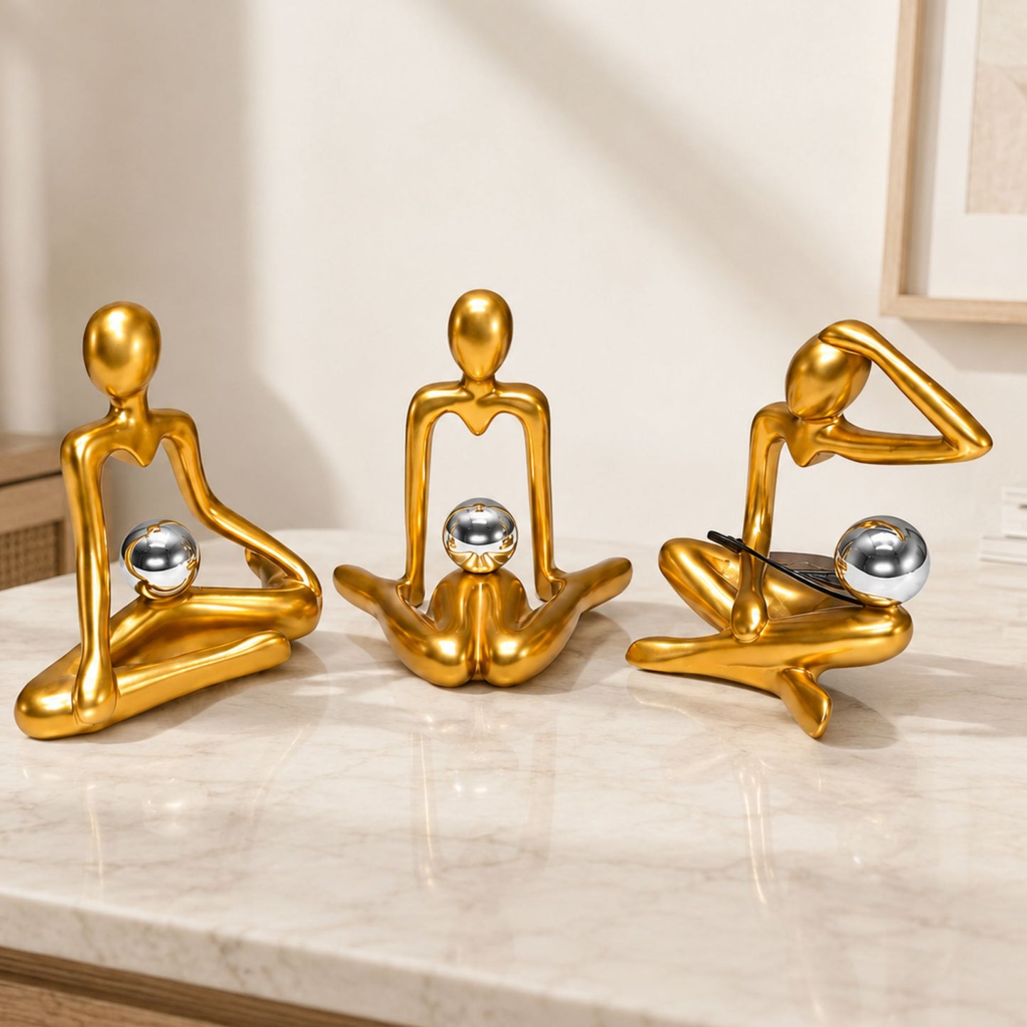 Set of three glossy gold abstract human figurines in yoga poses holding silver spheres for modern table decor.