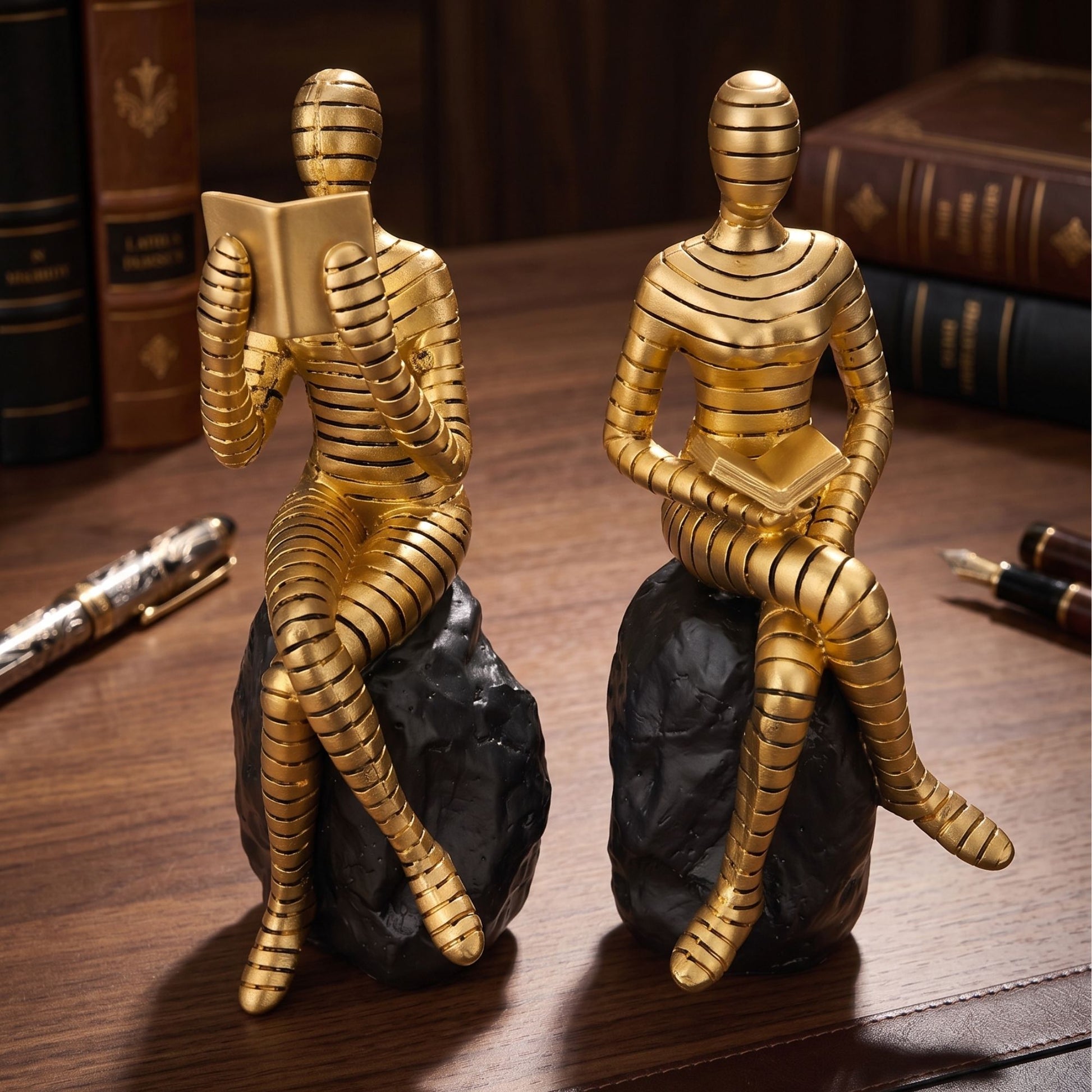 Set of two abstract metallic gold human figures reading books while sitting on black rocks for bookshelf and desk decor.
