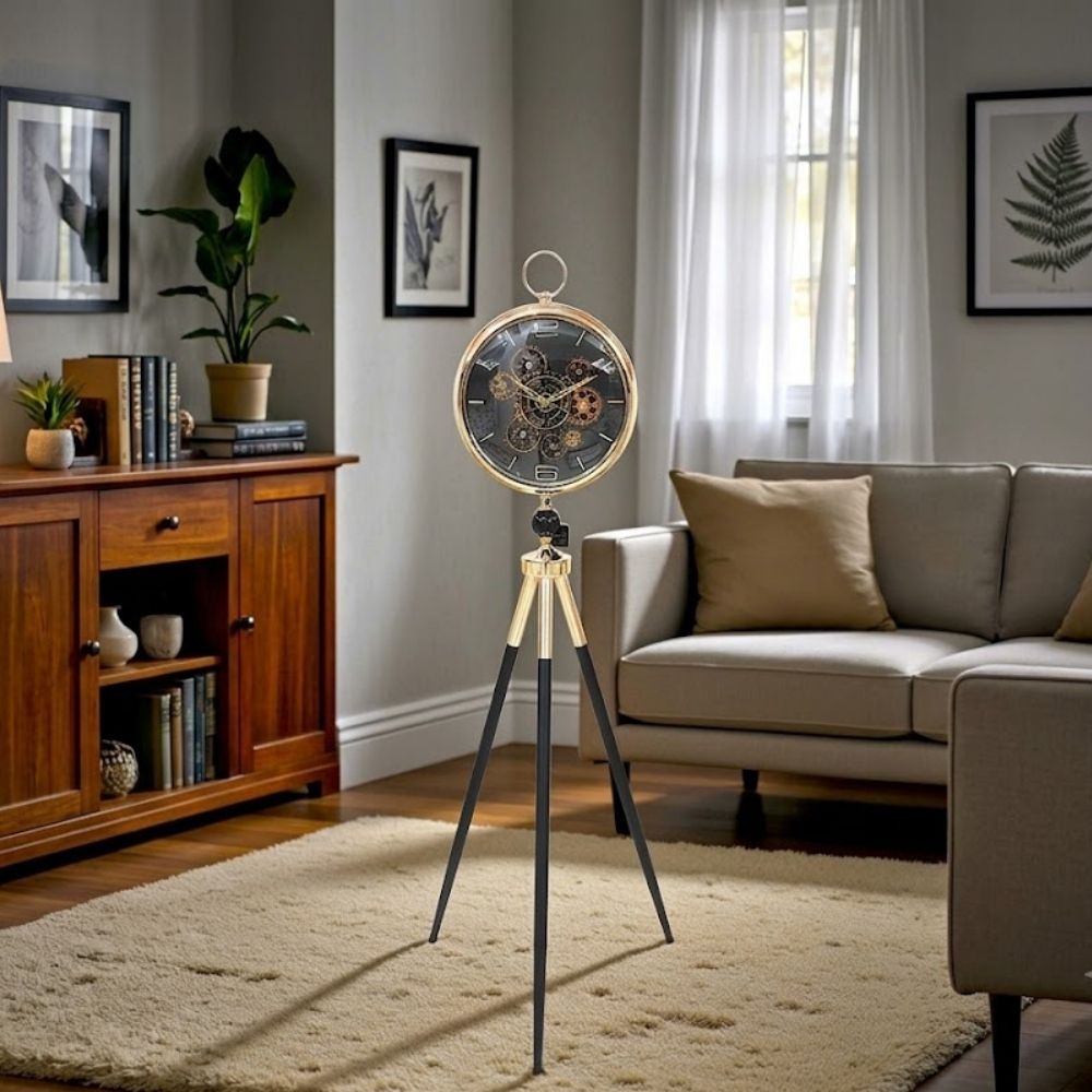 Standing tripod floor clock with slender black and gold legs, featuring a round gold-rimmed clock face with visible internal gears set against a dark dial in a modern living room setting.